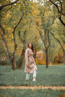 Pembe ceketli ve delikleri olan mavi kot pantolonlu bir kız sonbahar parkında yürüyor.