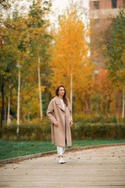 Pembe ceketli ve delikleri olan mavi kot pantolonlu bir kız sonbahar parkında yürüyor.