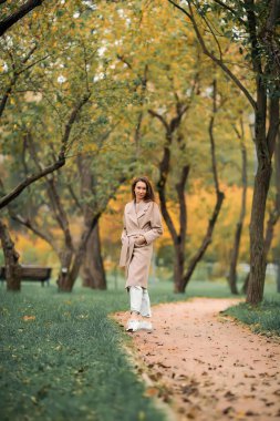 Pembe ceketli ve delikleri olan kot pantolonlu Avrupalı bir kız sonbahar parkında yürüyor.