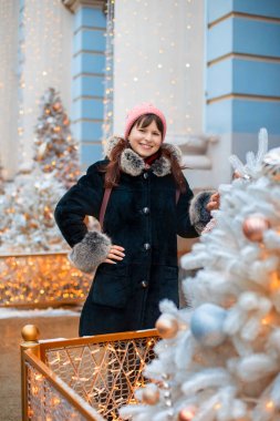 Siyah koyun derisi ceketli, kürklü ve pembe şapkalı bir kız süslü Noel ağaçlarının yanında poz veriyor.