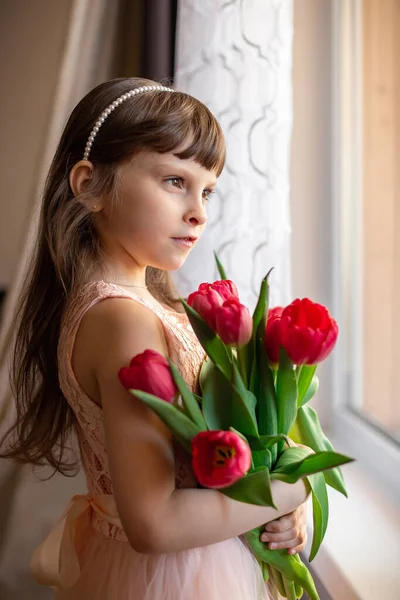 A Caucasian six-year-old girl in a pink dress holds red tulips in her hands and stands at the window