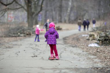 Pembe ve mor elbiseli dört yaşında beyaz bir kız parkta kırmızı bir scooter kullanıyor.