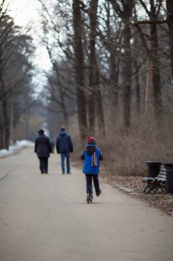 Avrupalı altı yaşındaki mavi elbiseli bir çocuk parkta scooter sürüyor.