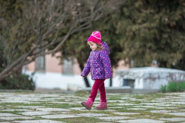 Mor ve pembe elbiseli dört yaşında beyaz bir kız parkta yürüyor.