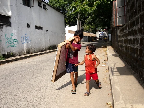 A young boy carries a piece of wood as his younger companion looks on and talks to him. — Stock Photo, Image