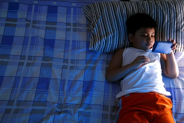 Niño jugando con un teléfono celular o smartphone en una cama —  Fotos de Stock