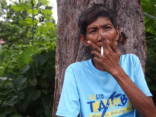 Um homem fuma um cigarro enquanto descansa e se inclina contra um tronco de árvore . — Fotografia de Stock