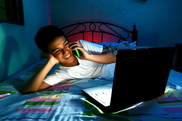 Ung tonåring framför en bärbar dator och på en säng och använder en mobiltelefon eller smartphone — Stockfoto
