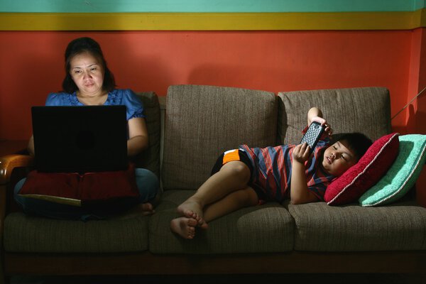 Woman using a laptop computer and a Young kid using a smartphone on a couch