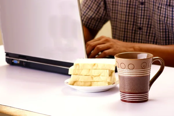 Tazza di caffè, pila di pane e uomo che lavora su un computer portatile in background — Foto Stock