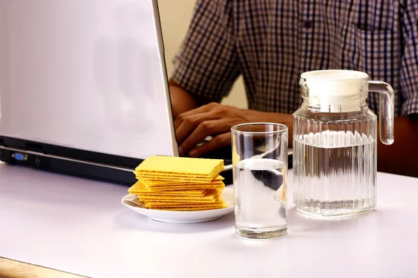 Bicchiere e brocca d'acqua e una pila di cracker e uomo che lavorano su un computer portatile in background — Foto Stock