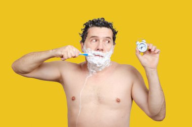 Ağzında diş fırçası olan bir adam, kafasını yıkıyor, tıraş oluyor ve çalar saate bakıyor.