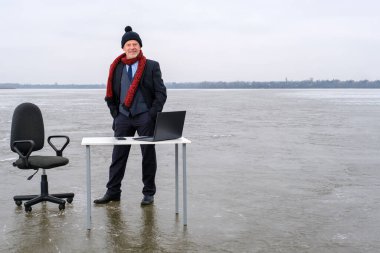 Takım elbiseli işadamı donmuş gölün ortasında bir masa ve sandalyede duruyor.