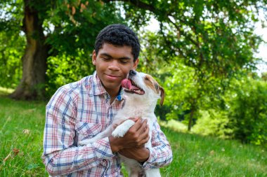 Genç Afro-Amerikan bir adam yazın Jack Russell teriyer köpeğini parkta tutuyor. Yazın parkta yürürken, alçak açılı bir manzara.