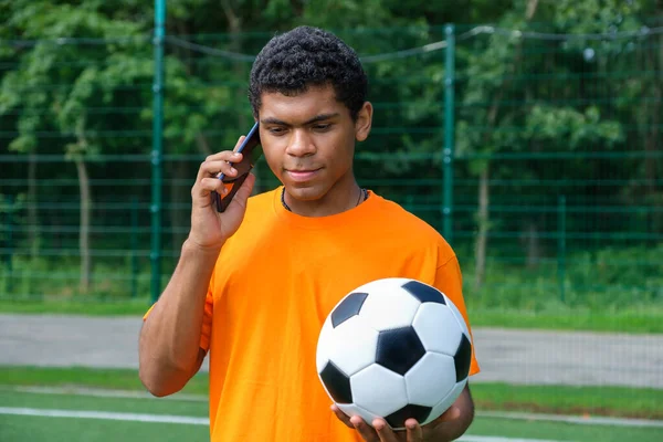 Brazilský muž drží fotbalový míč a používat smartphone na sportovním hřišti — Stock fotografie