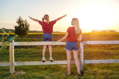 Genç anne ve kızı, gün batımında çitin yanında kollarını açmış, tarlada bekliyorlar. Kırsalda yürüyüş, kırsal alan, çiftçilik..