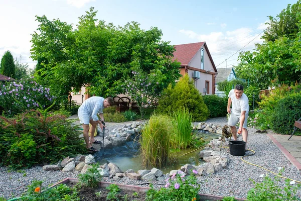 Olgun bir baba ve genç bir oğul suni bahçe havuzu dibini yüksek basınçlı çamur ve çamur hortumuyla temizliyor. Bahar ve yaz gölet bakımı