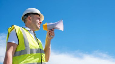 Beyaz miğferli bir miğfer içindeki mimar mavi gökyüzü arka planına karşı bir megafona bağırıyor.