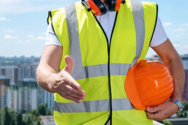 Budowniczy wyciąga jedną rękę na uścisk dłoni, a drugą trzyma pomarańczowy kask. — Zdjęcie stockowe