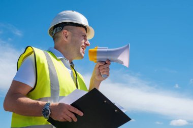 Beyaz miğferli bir miğfer içindeki mimar mavi gökyüzü arka planına karşı bir megafona bağırıyor.