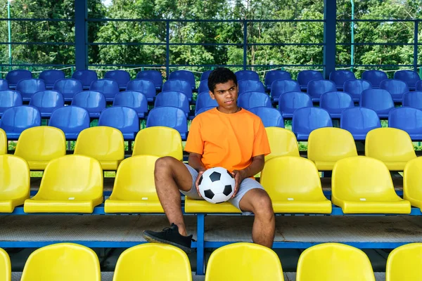 African American guy with a soccer ball sitting on an empty football grandstand — Stock Photo, Image