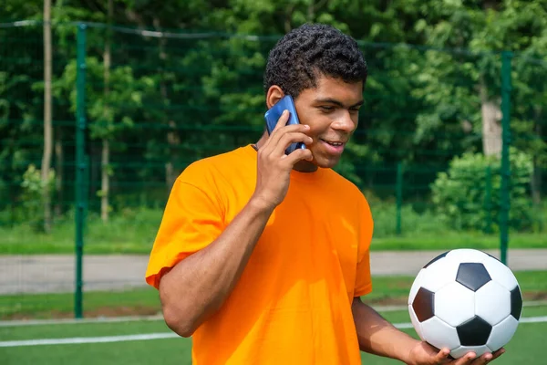 Brazilský muž drží fotbalový míč a používat smartphone na sportovním hřišti — Stock fotografie