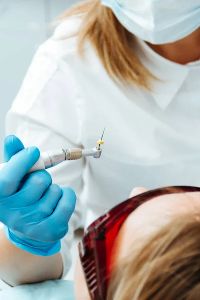 O dentista segura uma ferramenta de perfuração em luvas azuis em sua mão, sobre o paciente — Fotografia de Stock