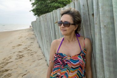 50 yaşında bir kadın. Bulutlu bir günde kumsalda, tahta bir çite yaslanmış.