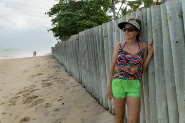 50 yaşında bir kadın. Bulutlu bir günde kumsalda, tahta bir çite yaslanmış.