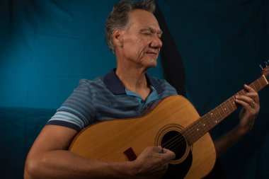 Yaşlı adam akustik gitarını mavi ışıklı arka planda çalarken eğleniyor.