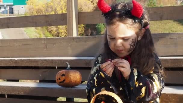 Een meisje in een heksenpak en een zwarte hoed op het hoofd met zwarte Halloween make-up, zombie die snoepjes eet bij een viering van Halloween — Stockvideo