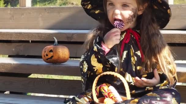 Uma menina em um terno de bruxa e um chapéu preto na cabeça com maquiagem de Halloween preto, zumbi comendo doces vermes de geleia em uma celebração do Dia das Bruxas — Vídeo de Stock