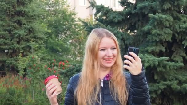Girl walks in the park with big purchases after shopping, talking on the phone. Happy girl using a smart phone in a city park sitting on a bench, drinks coffee, is happy, upset, surprised — Video Stock