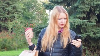 girl walks in the park with big purchases after shopping, talking on the phone. Happy girl using a smart phone in a city park sitting on a bench, drinks coffee, is happy, upset, surprised
