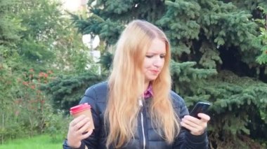 girl walks in the park with big purchases after shopping, talking on the phone. Happy girl using a smart phone in a city park sitting on a bench, drinks coffee, is happy, upset, surprised