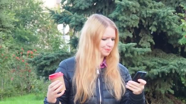 Дівчина гуляє в парку з великими покупками після покупки, розмовляючи по телефону. Щаслива дівчина, використовуючи смартфон у міському парку, сидить на лавці, п'є каву, щаслива, засмучена, здивована — стокове відео