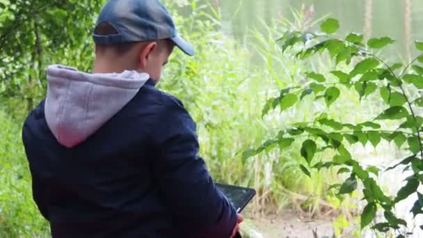 Mignon garçon enfant jouer avec tablette PC de plein air avec forêt sur fond près du lac jeu informatique concept de dépendance — Video