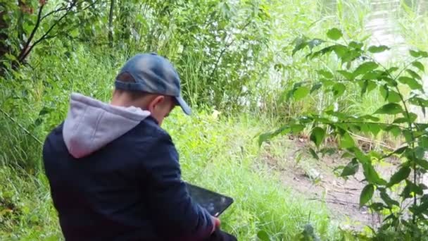Mignon garçon enfant jouer avec tablette PC de plein air avec forêt sur fond près du lac jeu informatique concept de dépendance — Video