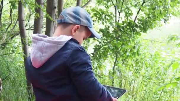 Mignon garçon enfant jouer avec tablette PC de plein air avec forêt sur fond près du lac jeu informatique concept de dépendance — Video