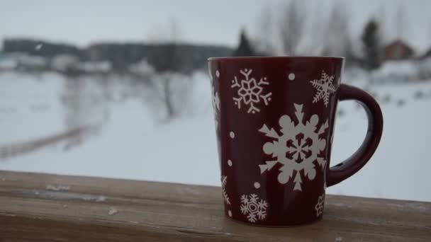 Piros vagy burgundi csésze mintázatú hópelyhek a háttérben a téli erdőben. havazik. Újév. Karácsonykor. csésze forró itallal az ablakpárkányon egy téli város hátterében. — Stock videók