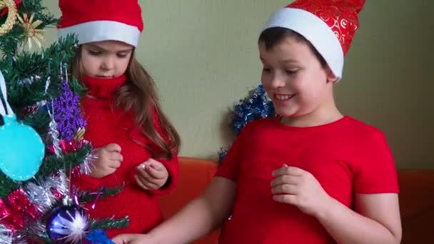 Cute little brother and sister celebrate christmas or new year at home. Dressed in Christmas hats. The beautiful children decorating a Christmas Tree. joy fun, laugh — Wideo stockowe