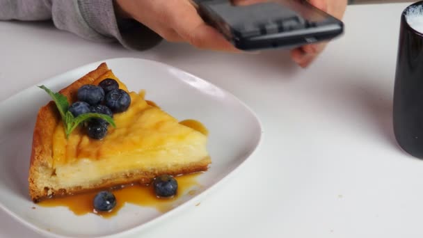Närbild flicka äter krämig mascarpone ost tårta blåbär i ett café med kaffe, med telefon, skriva ett meddelande, titta på nyheter, sociala nätverk. Cheesecake från New York. Juldessert. Hälsosam mat. — Stockvideo