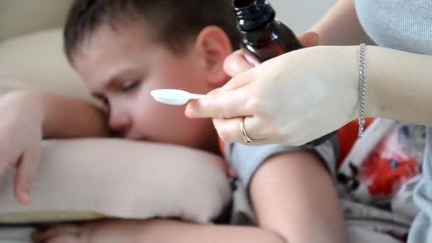 Le garçon est couché à la maison avec une fièvre, avec une toux, est traitée. Enfant malade garçon couché au lit, se reposant à la maison maman verse de la potion dans une cuillère — Video