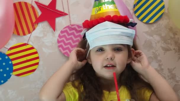 Portret pięknej dziewczynki z tortem urodzinowym. dziewczyna w czapce świętuje swoje urodziny, bawi się, dmucha fajkę, zdmuchuje świece na torcie, zjada ciasto, pije sok, nosi maskę podczas — Wideo stockowe