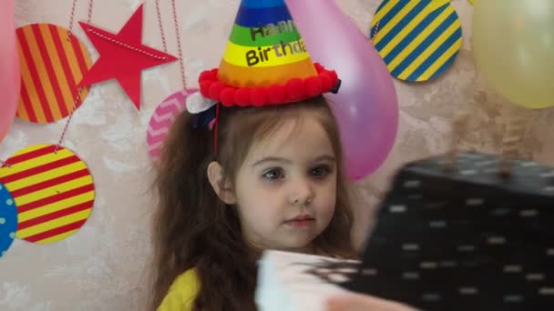 Portrét malé hezké holčičky s narozeninovým dortem. dívka v čepici slaví své narozeniny, baví, fouká dýmku, sfoukne svíčky na dortu, jí dort, pije džus, nosí masku během — Stock video