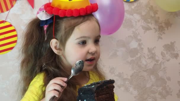 Ritratto di bambina carina con torta di compleanno. una ragazza con un cappello festeggia il suo compleanno, si diverte, soffia un tubo, soffia le candele sulla torta, mangia una torta, beve succo, indossa una maschera durante un — Video Stock
