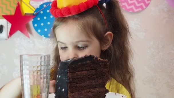 Doğum günü pastası ile küçük güzel bir kızın portresi. Şapkalı bir kız doğum gününü kutlar, eğlenir, pipo üfler, pastadaki mumları söndürür, pasta yer, meyve suyu içer, maske takar. — Stok video