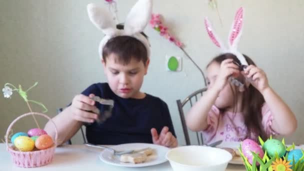 Jongen en meisje bereiden koekjes voor Pasen, lachen. Plezier hebben op de paaseieren jacht. Kind en meisje met konijnenoren en eieren aan. kleurrijke eieren. — Stockvideo