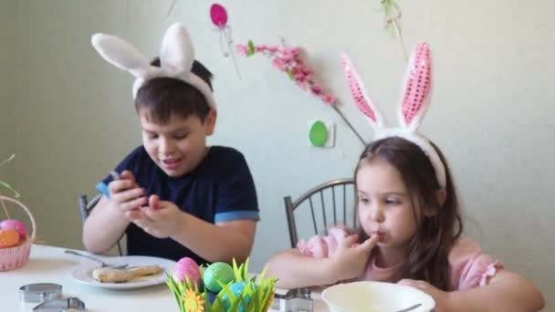 Chłopiec i dziewczynka przygotowują ciasteczka na Wielkanoc, śmieją się. Dobrze się bawisz podczas polowania na pisanki. Chłopiec i dziewczynka noszą królicze uszy i malują jajka. kolorowe jaja. — Wideo stockowe