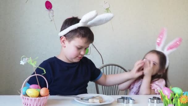 Chłopiec i dziewczynka przygotowują ciasteczka na Wielkanoc, śmieją się. Dobrze się bawisz podczas polowania na pisanki. Chłopiec i dziewczynka noszą królicze uszy i malują jajka. kolorowe jaja. — Wideo stockowe